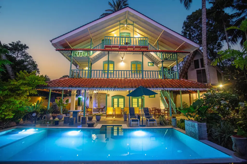 Sekku House at dusk: a two-storey colonial-style villa with timber balconies, turquoise shutters and a lit pool in front